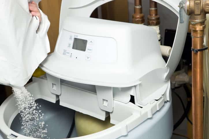 A person pours white salt crystals from a bag into the open brine tank of a home water softener unit during routine maintenance.